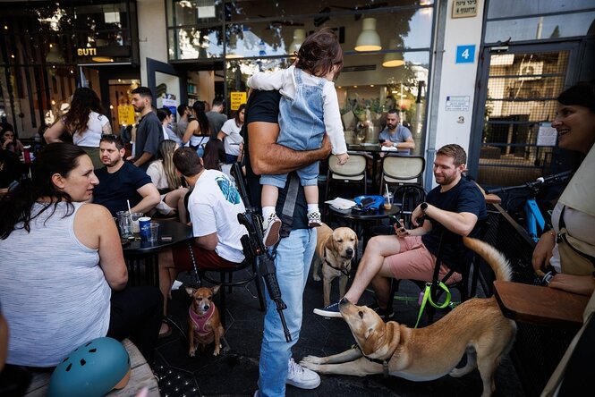 Menschen in einem Café. Ein Mann trägt ein Gewehr, er hat ein Kind auf dem Arm