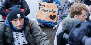 Aktivist*innen der letzten Generation bei einer Blockadeaktion Mitte März. "Ganz ehrlich? Fliegen oder Überleben" steht auf einem Plakat, das ein Klimaaktivist hält.Bundesweit finden Demonstrationen der Letzten Generation mit Ärzten, Wissenschaftlern, Handwerkern, Studenten und Rentnern Statt.