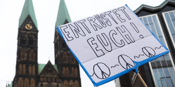 Ein Demonstrant hält während des Ostermarsches 2018 vor dem Dom und der Bürgerschaft ein Schild mit der Aufschrift "ENTRÜSTET EUCH!" nach oben.