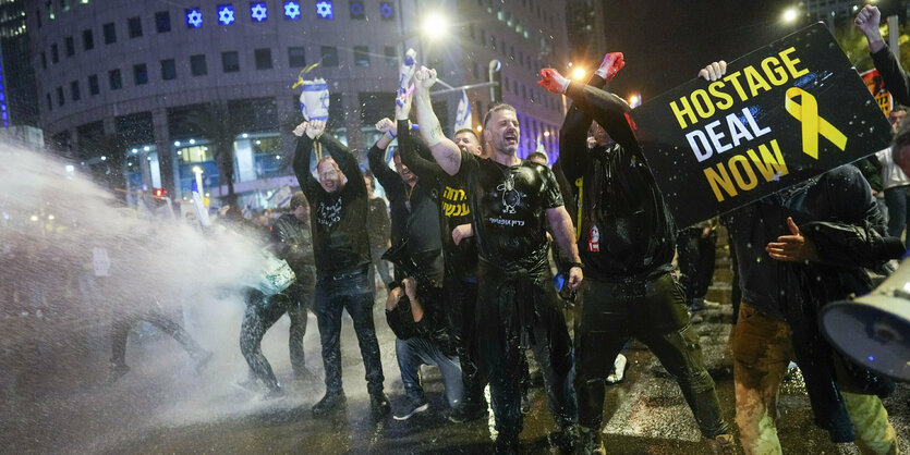 Menschen halten Protestplakate, hinter ihnen wird Tränengas eingesetzt