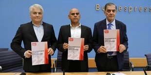 Dr. Mehmet Daimagüler, Dr. Guillermo Ruiz Torres und Romani Rose stehen zusammen in der Bundespressekonferenz in Berlin und halten den Bericht in die Kamera