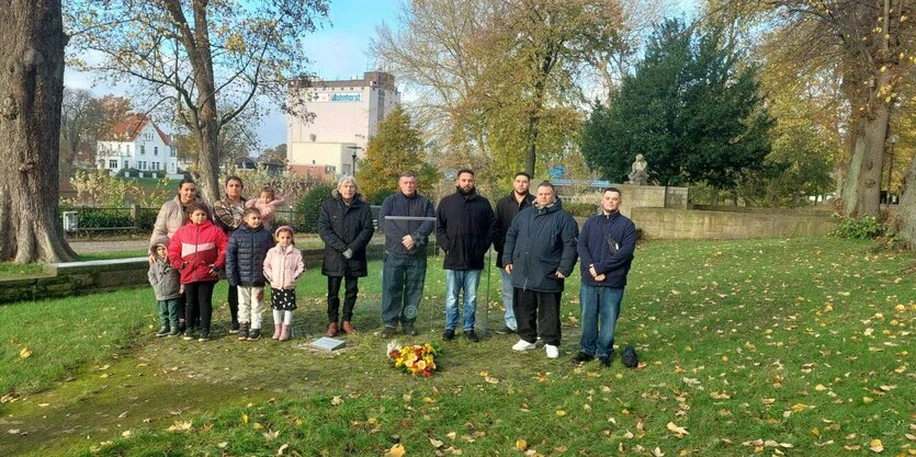 Eine Gruppe Menschen, Männer, Frauen und Kinder, steht vor einer Gedenktafel mit einem Blumenstrauß