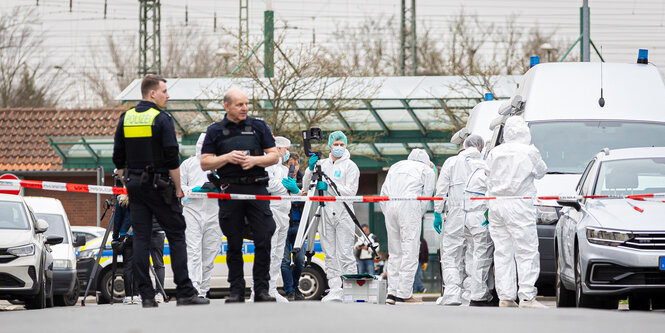 Hinter einer Absperrung stehen Menschen in Schutzanzügen und Polizisten in Uniform