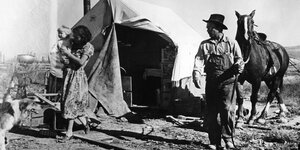 Eine Frau mit Kind auf dem Arm vor einem Zelt, daneben ein Mann in ARbeitskleidung mit Pferd, historische Aufnahme