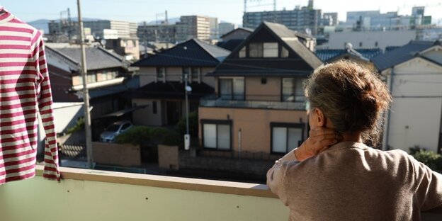 Blick von einem Balkon. Eine ältere Frau steht mit dem Rücken zur Kamera und blick von ihrem Balkon aus auf eine Stadt. Sie hat das Gesicht auf die linnke Hand gestützt. Links im Bild hängt ein rot-weiß gestreiftes Hemd auf einem Bügel zum Trocknen in der