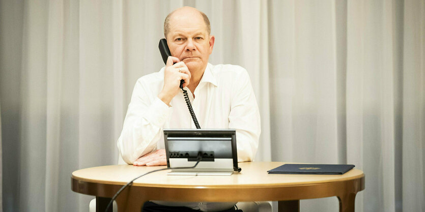 Olaf Scholz sitzt an einem kleinen ovalen Tisch vor einem hellen Vorhang, er hält den Telefonhörer ans Ohr und schaut ernst in die Kamera