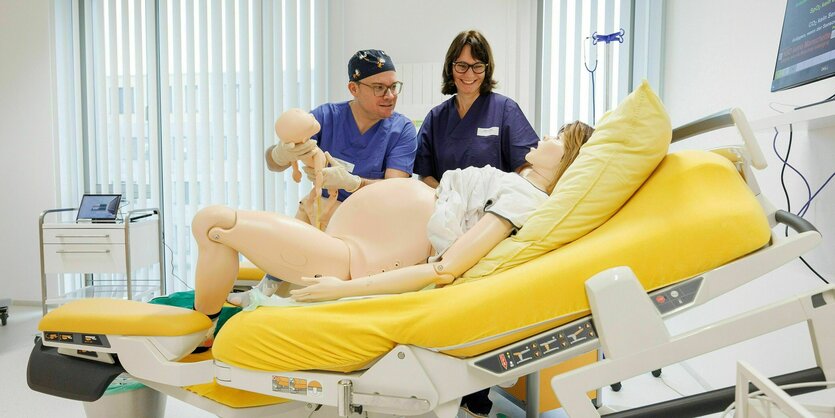 Eine Hebamme und ihr Auszubildender stehen in einem Simualtionslabor, auf dem Bett liegt der Dummy einer Gebärdenen. Der Auszubildende hält ein Dummy-Baby hoch