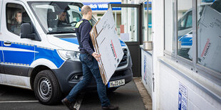 Ein Polizei-Bulli steht vor einem Gebäude, ein uniformierter Polizist trägt einen gefalteten Karton hinein.