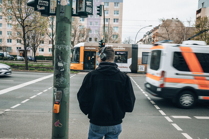 Ein mann steht an einer Ampelkrezung und blickt auf den Verkehr. Er trägt Ohrenschützer