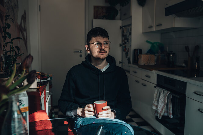 Ein mann sitzt in seiner Küche, schaut nachdenklich und hält eine rote Tasse in den Händen