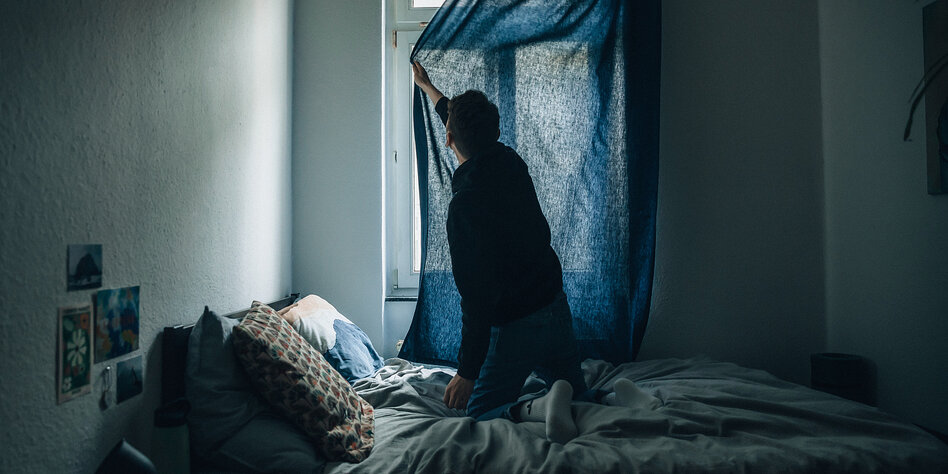 Ein Mann hockt auf seinem Bett und zieht einen dunkelblauen Vorhang vor das Fenster in seinem Zimmer