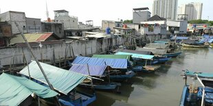 Bote liegen in einem Kanal, die Häuser am Ufer sind durch eine Mauer geschützt, im Hintergrund sind zwei Hochhäuser zu sehen