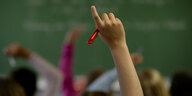 Ein Kind meldet sich im Unterricht. man sieht nur den nackten Unterarm und die Hand, die gleichzeitig einen roten Stift hält. Im Hintergrund andere Kinde rund eine grüne Tafel.