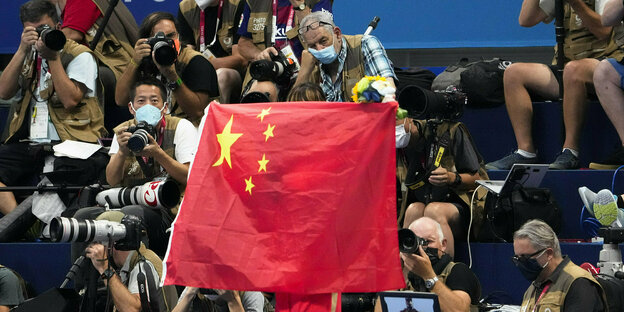 Eine Person hält eine chinesische Flagge mit dem Rücken zur Kamera, vor der Personen stehen und sitzen Kamerateams