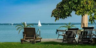 Leere Holz-Liegestühle stehen am Ufer des Chiemsees auf dem Segelboote unterwegs sind