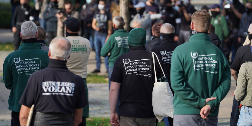 Eine kleine Gruppe von Männern steht mit dem Rücken zur Kamera, auf ihren Pullovern stehen Aufschriften wie "National Revolutionär Sozialistisch" und "Deutsche Jugend voran!"