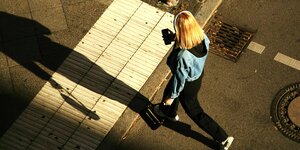 Eine junge Frau traegt Kopfhoerer und blickt auf ihr Smartphone, während sie über eine Straße geht