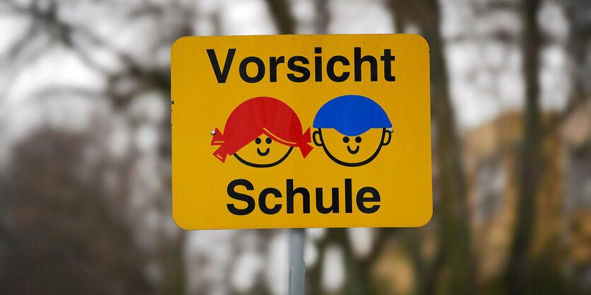 Ein Schild steht vor Bäumen das zwei gezeichnete SchülerInnen zeigt und dem Hinweis: Vorsicht Schule