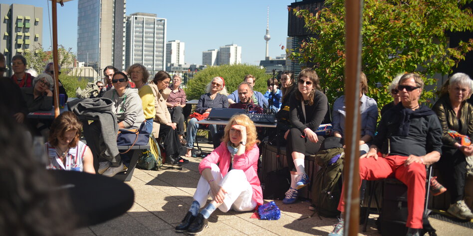 Menschen sitzen auf einer sonnigen Dachterasse auf Stühlen und auf dem Boden. Im Hintergrund die "Skyline" von Berlin mit dem Fernsehturm.