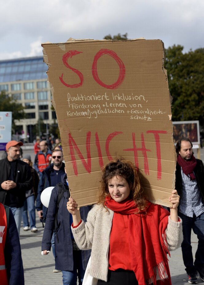 Eine Frau mit einem roten Schal trägt ein Pappschild auf den Schultern, darauf steht: "So nicht"