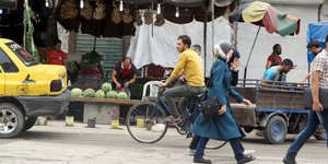 Menschen laufen auf der Straße und fahren Fahrrad, im Hintergrund Marktstände
