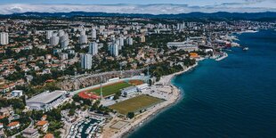 Vogelperspektive auf ein Stadion am Meer, darum herum Häuser.