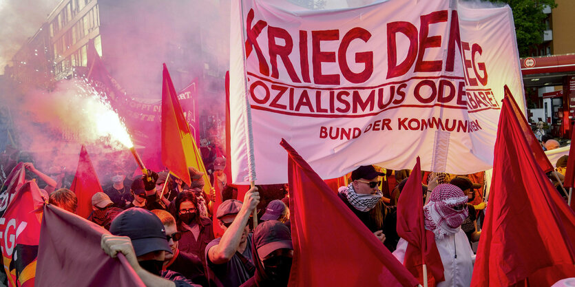 Vermummte Menschen halten ein Banner mit der Aufschrift "Krieg dem Krieg. Sozialismus oder Barbarei". Rundherum tragen Leute rote Fahnen, jemand zündet ein Bengalo.