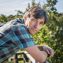 Foto von Ronald Prieß; er trägt ein blau kariertes Holzfällerhemd, dreht seinen Kopf von der Seite in Richtung Kamera und stützt seine Arme dabei auf ein Balkongeländer