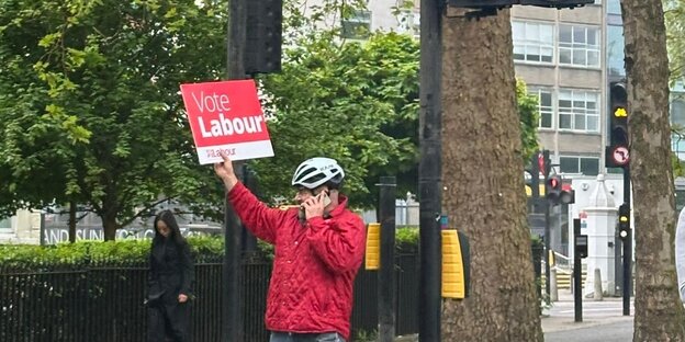 Eine Person mit Helm hält ein Schild mit der Aufschrift „Vote Labour“ hoch