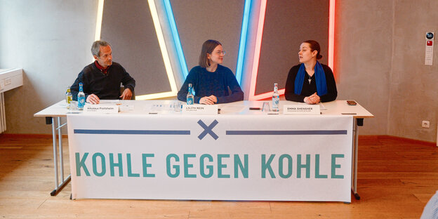 Drei Personen sitzen bei einer Pressekonferenz an einem Tisch.  Davor hängt ein Plakat mit dem Slogan: Kohle gegen Kohle