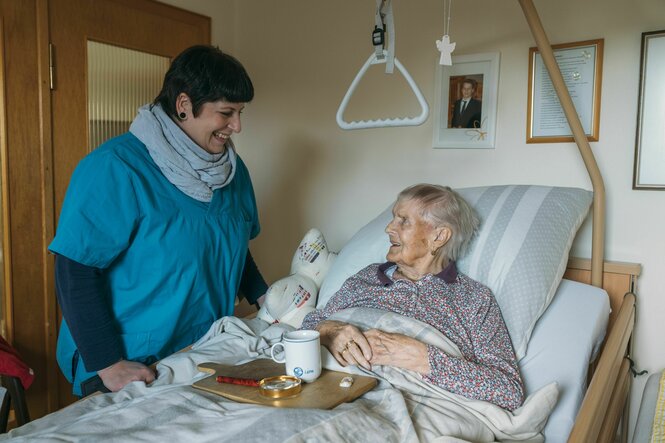 Eine Mitarbeiterin des Pflegedienstes steht am Bett einer Kundin