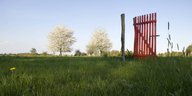 Ein rotes Gittertor steht auf einer Rasenfläche, im Hintergrund Bäume