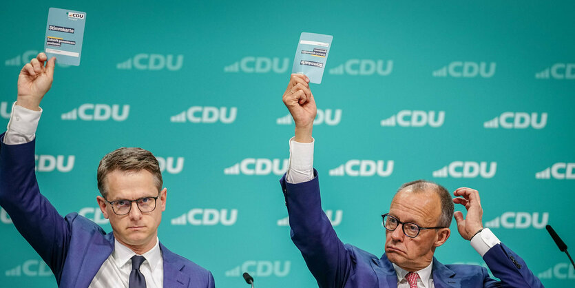 Carsten Linnemann und Friedrich Merz sitzen auf einem Podium und heben ihre Stimmkarten