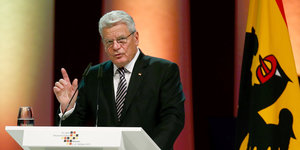 Mann mit weißen Haaren (Bundespräsident Gauck) hebt Finger vor Deutschlandfahne