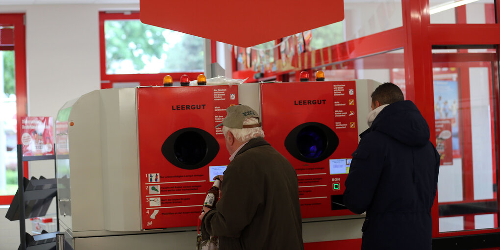 Zwei Männer stecken Pfandflaschen in einen Pfandflaschenautomaten in einem Supermarkt.