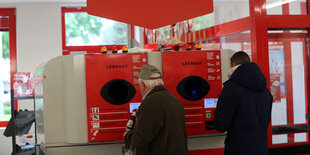 Zwei Männer stecken Pfandflaschen in einen Pfandflaschenautomaten in einem Supermarkt.