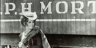 Filmstill aus dem Schwarz-Weiß-Film „Spiel mir das Lied vom Tod“ (1968). Claudia Cardinale steht vor einem Zugwaggong, auf dem Teile einer Aufschhrift zu lesen sind: "P. H. Mort". Sie trägt einen flachen Cowboyhut und hat ein Tuch über die Schulter geworfen. Sie lächelt und schaut nach rechts.