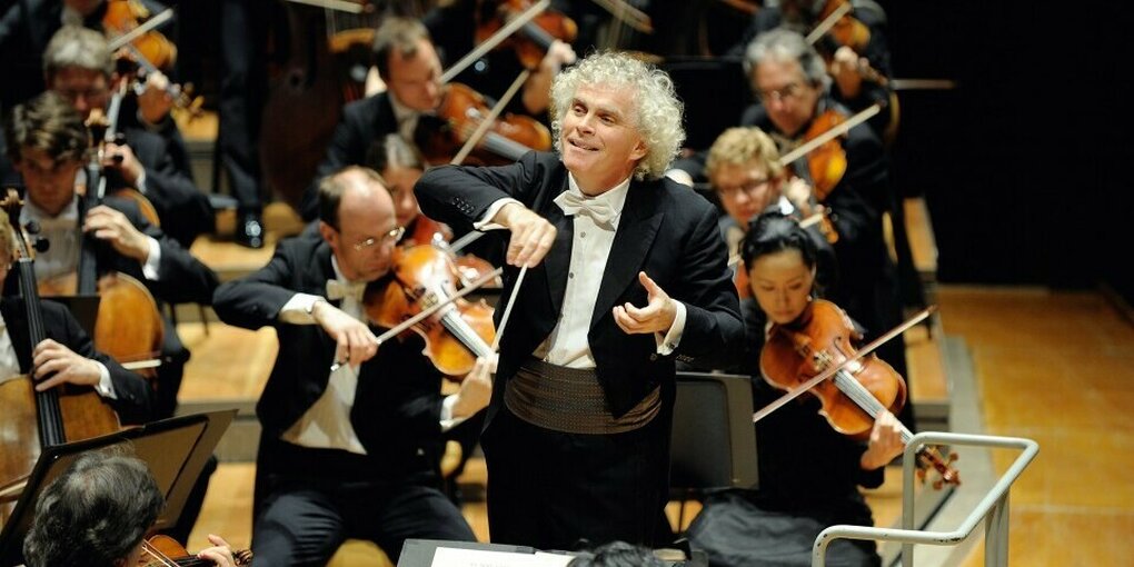 Blick auf ein Orchester. Der Dirigent Simon Rattle steht am Dirigentenpult und bewegt den Taktstock und seine Hand zur Musik. Er lächelt.