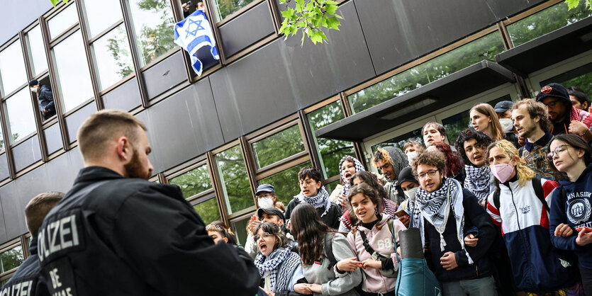 Zwei Polizisten stehen einer Gruppe junger Menschen gegenüber, die Kufijas tragen. Aus einem Fenster über den Leuten hängt eine Israelfahne.