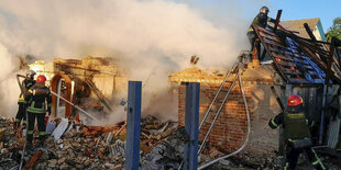 Feuerwehrleute und Rettungskräfte vor und in Trümmern eines Hauses. Es steigt Rauch auf. Im Hintergrund ein klein wenig blauer Himmel.