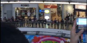 Ein Videostill das die singenden Protestierenden in einer Shopping Mall zeigt.