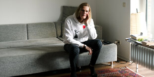 Greta Bollig sitzt in Leggins und einem grauen Shirt mit Bansky-Motiv auf einem grauen Sofa, vor dem ein rot-gemusterter Teppich liegt. Licht fällt seitlich durchs Fenster.