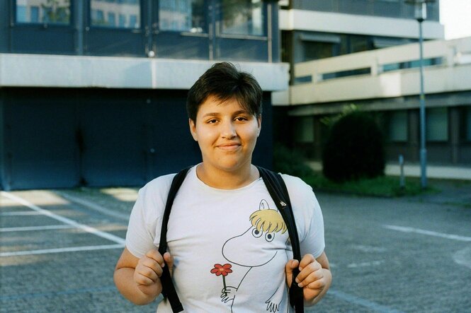 Maaradji steht auf einem Parkplatz vor einer Brutalo-Beton-Architektur, er trägt einen Rücksack, ein T-shirt mit lachendem Pferd und lächelt