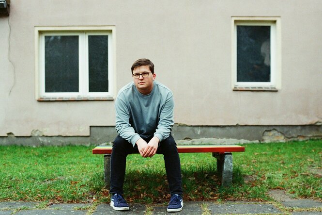 Marius sitzt vor einem Haus auf einer Bank, die auf einem Grünstreifen steht, er trägt eine Brille und einen hellen Pullover