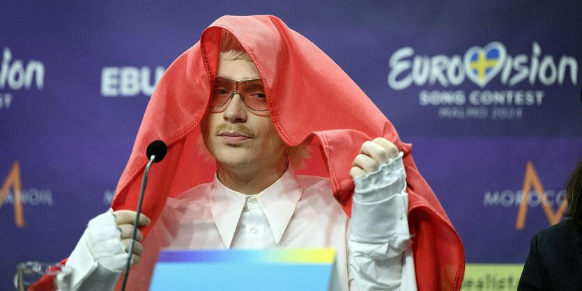 Joost Klein in weißen Klamotten sitzt bei einer Pressekonferenz vor einem Mikro und bedeckt sein Haupt mit einer roten Textilie