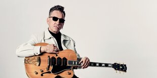 Richard Hawley mit dunkler Sonnenbrille, weißer Jeansjacke und Gitarre im Arm