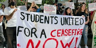 Eine Gruppe von Menschen steht unter einem Baum, viele tragen karierte Tücher - Kufijas. Sie halten große Banner, auf ihnen steht "Students United for a Ceasefire Now" und "your silence is complicity"