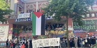 Rund 30 vermummte Personen stehen vor einerm graffitibesprühten Haus und halten Transparente und eine Palästina-Flagge