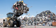 Ein Bagger transportiert auf einer Müllhalde eine große Schaufel Elektroschrott, im Hintergrund liegt ein weiterer Schrottberg