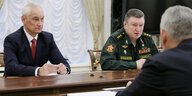 Andrei Belousov und Alexander Lapin sitzen zusammen an einem Tisch, ihnen gegenüber ein Mann, der nicht zu erkennen ist - sie schauen ernst